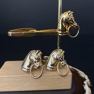 Op de afbeelding: Goudkleurige metalen paardenhoofdmanchetknopen en stropdasklemset. De paardenkoppen hebben gedetailleerde manen en een ring in de mond. De set wordt gepresenteerd op een houten blok en een gouden standaard.