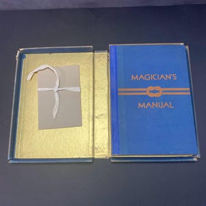 Puede incluir: Un libro azul titulado Magician's Manual con texto naranja y un gráfico de nudo en la portada, alojado en una caja forrada en oro. Una tarjeta rectangular beige atada con un cordel blanco está sujeta al lado interior izquierdo de la caja abierta.