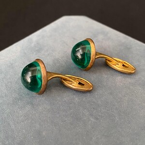 Könnte beinhalten: Ein Paar Vintage-Manschettenknöpfe mit goldfarbenem Metallrahmen. Jeder Manschettenknopf hat einen abgerundeten, durchscheinenden, smaragdgrünen Stein. Die Manschettenknöpfe werden auf einer hellblauen Oberfläche präsentiert.