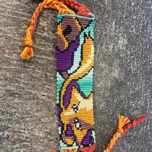 Peut inclure: Un bracelet d'amiti&eacute; color&eacute; avec un motif tiss&eacute; repr&eacute;sentant un personnage de dessin anim&eacute;. Le bracelet est r&eacute;alis&eacute; avec des fils orange, jaune, vert, violet et marron. Le bracelet a de longues franges orange et vertes.