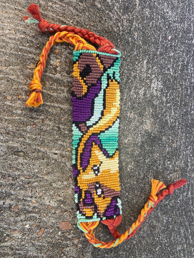 Peut inclure: Un bracelet d'amiti&eacute; color&eacute; avec un motif tiss&eacute; repr&eacute;sentant un personnage de dessin anim&eacute;. Le bracelet est r&eacute;alis&eacute; avec des fils orange, jaune, vert, violet et marron. Le bracelet a de longues franges orange et vertes.