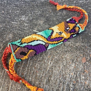 Peut inclure: Un bracelet d'amiti&eacute; color&eacute; avec un motif pixelis&eacute; d'un personnage de dessin anim&eacute;. Le bracelet est fait de fils orange, jaune, vert, violet et marron.