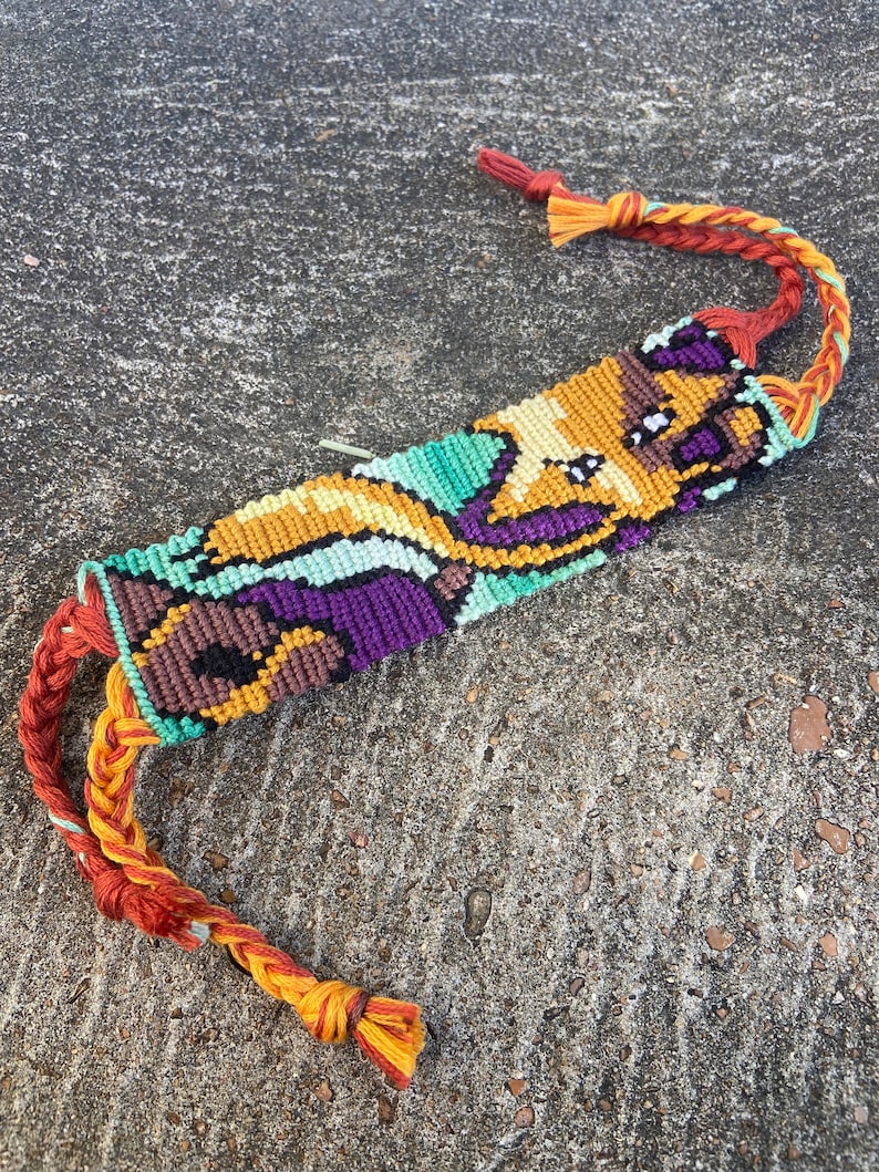 Peut inclure: Un bracelet d'amiti&eacute; color&eacute; avec un motif pixelis&eacute; d'un personnage de dessin anim&eacute;. Le bracelet est fait de fils orange, jaune, vert, violet et marron.