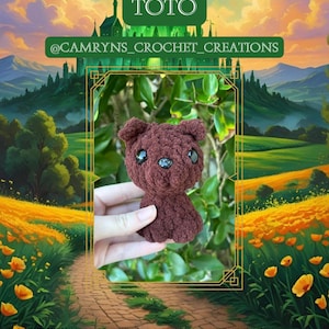 May include: A crocheted brown bear with black button eyes is held in front of a painted landscape. The bear is labeled "TOTO" and the seller's name is displayed above the bear. The background features a path, flowers, and a castle.