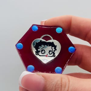 May include: A hexagonal, burgundy-colored charm with a silver heart-shaped emblem featuring a cartoon character. The charm has six blue dots around the edges. The charm is held in a hand.