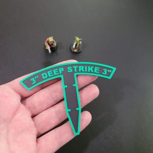 May include: Black and green 3" deep strike marker for tabletop games. The marker is shaped like a T with a rounded top and a pointed bottom. The text "3" DEEP STRIKE 3" is printed in green on the top of the marker.
