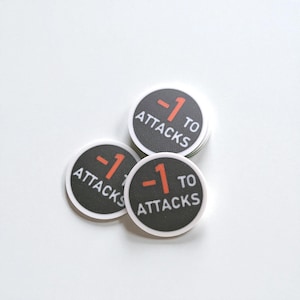 May include: A stack of black and white game tokens with the text "-1 TO ATTACKS" in white and red. The tokens are circular and have a white border. The background is white.