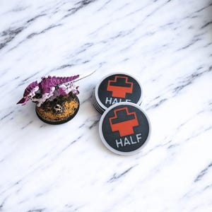 May include: A miniature purple and white creature figurine on a textured base, next to a stack of black and white circular tokens. The tokens have a red cross symbol and the word "HALF" printed on them. The background is a white marble pattern.