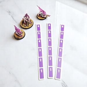 May include: Miniature purple creature figurines with white detailing on circular bases, with three purple and white numbered markers. The markers are numbered from 1 to 6. The bases are brown and yellow.