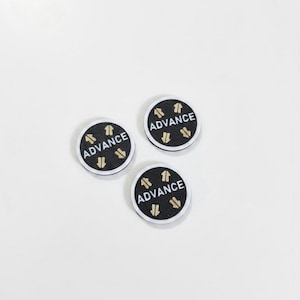 May include: Three circular buttons with a black textured surface, a silver-toned metallic edge, and the word ADVANCE printed in white capital letters, accompanied by four small gold-colored arrow icons pointing in alternating directions.