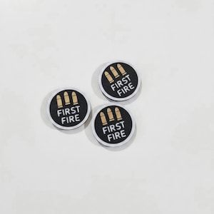 May include: Three circular black patches with a silver border. Each patch features a graphic of three gold-colored bullets arranged horizontally above the white text FIRST FIRE, printed in a bold, sans-serif font centered on the black background.