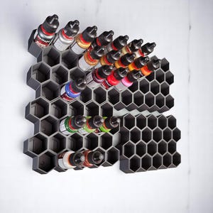 May include: A grey honeycomb-shaped wall organiser with multiple rows of small bottles of paint. The bottles are arranged in a grid pattern and are of various colours, including red, blue, green, yellow, and black. The organiser is made of plastic and has a textured surface.