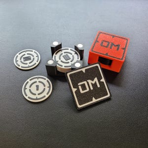 May include: A black and white game piece with the letters "DM" on it. The game piece is sitting on a black surface. There are also several black and white game pieces in a stack, and a red box with the letters "DM" on it.