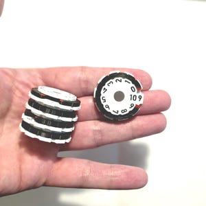 Puede incluir: Una pila de cinco discos circulares numerados en blanco y negro junto a un disco individual a juego. Cada disco tiene un orificio central y números negros del cero al nueve dispuestos en círculo, con pequeños acentos rojos en el borde.