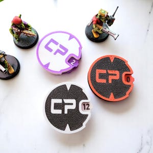 Op de afbeelding: Drie ronde, witte plastic tokens met zwarte tekst en cijfers. De tokens hebben de letters "CP" en een nummer op elk. De nummers zijn 2, 0 en 12. De tokens liggen op een wit oppervlak.