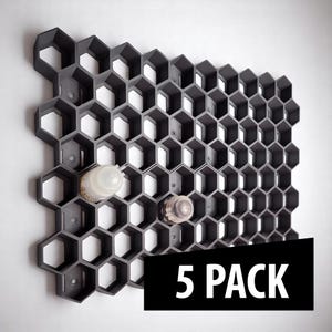 Op de afbeelding: Een donkergrijze, honingraatvormige wandorganizer met twee doorzichtige flessen in enkele van de cellen. De organizer is aan een witte muur bevestigd. De tekst "5 PACK" wordt in een zwarte rechthoek onderaan weergegeven.