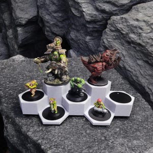 Puede incluir: Un expositor hexagonal blanco con seis plataformas para mostrar figuras en miniatura. Las figuras están pintadas en varios tonos de verde, rojo y marrón. Las figuras incluyen un gran ogro verde, un pequeño goblin verde y un demonio rojo.