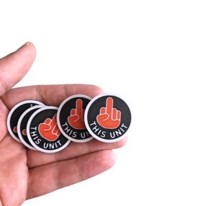 Könnte beinhalten: Eine Sammlung runder schwarz-weißer Aufkleber mit einer roten Handgeste und dem Text "THIS UNIT". Die Aufkleber werden in einer Hand vor weißem Hintergrund gehalten.