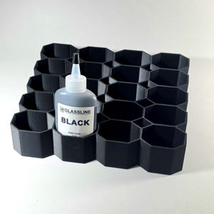 May include: A black 3D-printed organizer with a honeycomb grid pattern featuring multiple octagonal compartments. One compartment holds a squeeze bottle labeled GLASSLINE BLACK with a white cap and a pointed nozzle for precise application.