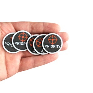 Puede incluir: Una mano sostiene varias fichas circulares en blanco y negro. Cada ficha tiene un borde blanco, la palabra "PRIOR" o "PRIORITY" en blanco y un símbolo de diana rojo. Fondo blanco.