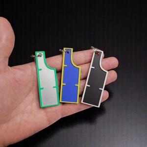 May include: Three keychains with different coloured backgrounds. The keychain on the left has a silver background with a green border. The keychain in the middle has a blue background with a yellow border. The keychain on the right has a black background with a white border.