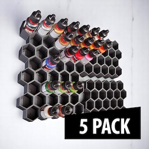 May include: A dark gray, honeycomb-shaped paint bottle organizer holding several bottles of paint in various colors. The organizer is mounted on a white surface. The text "5 PACK" is displayed in a black rectangle.