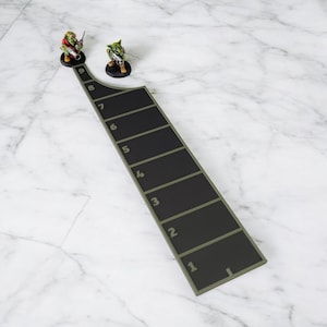 May include: A black and green game board with numbered sections, alongside two miniature goblin figures. The board has a curved design and is set against a marble background, suggesting a tabletop game setting.
