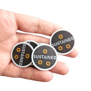 May include: A hand holding several small, round black tokens with white borders. Each token has the word "SUSTAINED" in white text and three small, yellow starburst designs. The tokens are likely game pieces or decorative items.