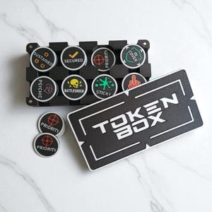May include: A black token box with the text "TOKEN BOX" in white. The box holds several round tokens with various symbols and text, including "SUSTAINED," "SECURED," and "PRIORITY." Additional loose tokens are also visible.