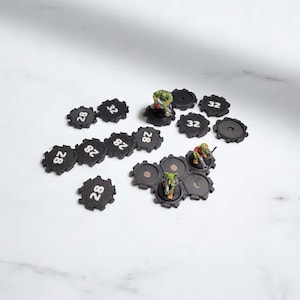 May include: A set of black plastic hexagonal game bases with white numbers, including 28 and 32. The bases are arranged in a pattern on a white surface. There are also two small green and red painted figures on the bases.