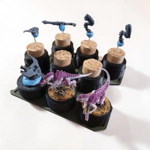 May include: A collection of miniature figurines and components, including purple and white creatures, gray armor pieces, and various other parts, all mounted on corks and bases. The items are arranged on a dark green tray.