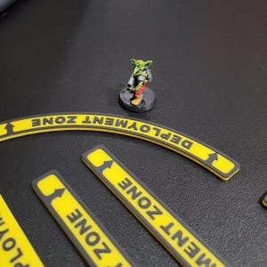 May include: A green and yellow miniature figure of a goblin holding a weapon stands on a black base. The figure is on a black surface with yellow and black "Deployment Zone" signs.