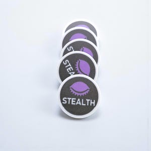 Pode incluir: Uma pilha de fichas redondas, pretas e brancas, com a palavra "STEALTH" impressa em branco. Cada ficha tem um gráfico roxo em forma de olho. As fichas estão dispostas em uma linha vertical sobre um fundo branco.
