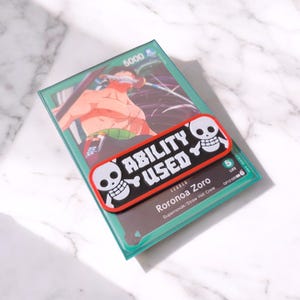 Peut inclure: Une carte à collectionner verte avec une illustration d'un personnage avec une épée. Un autocollant noir et rouge avec les mots "ABILITY USED" et des graphiques de crâne est posé sur la carte. La carte est intitulée "Roronoa Zoro".