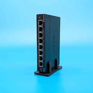 May include: A black Netgear network switch with multiple ports, standing upright on a black base. The device has the Netgear logo and other text visible on its surface. The background is a solid light blue.