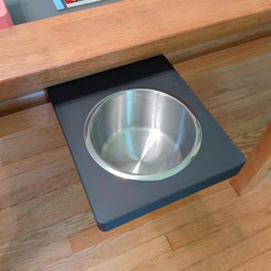 May include: A stainless steel cup holder insert mounted into a matte black rectangular platform, which is attached to the underside of a light-toned wooden table surface. The metal cup holder features a smooth, circular, reflective interior.