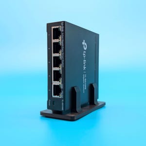 May include: A black TP-Link TL-SG105E 5-Port Gigabit Easy Smart Switch. The device is on a brown stand and has five numbered ports. The power button is visible on the lower left. The brand name and model are printed on the device.