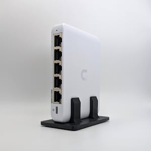 May include: A white network switch with multiple Ethernet ports, standing upright on a black stand. The device has a small, curved logo on its side. The ports are visible on the front, and the device is designed for network connectivity.
