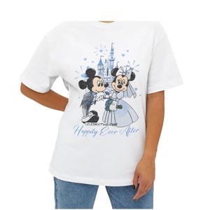May include: White t-shirt featuring a cartoon illustration of Mickey and Minnie Mouse in wedding attire, with a castle backdrop. The text "Celebrating Our Happily Ever After" is printed in blue.
