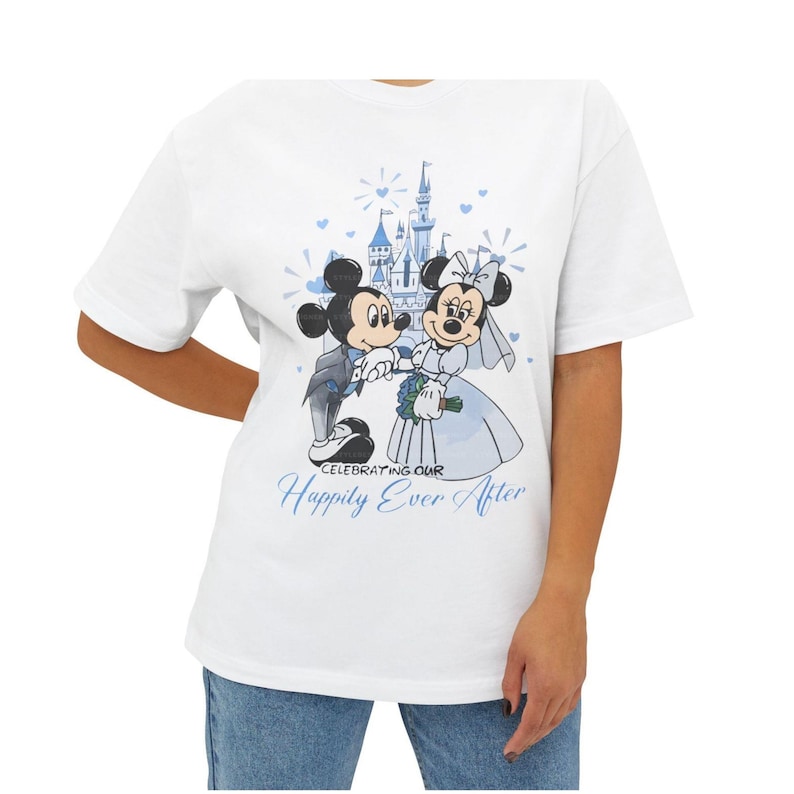 May include: White t-shirt featuring a cartoon illustration of Mickey and Minnie Mouse in wedding attire, with a castle backdrop. The text "Celebrating Our Happily Ever After" is printed in blue.