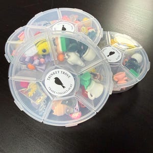 May include: Three clear, segmented containers filled with miniature trinkets. Each container has a label with a bird silhouette and the words "TRINKET TROVE, TRADE, COLLECT, PLAY." The trinkets include small figurines, beads, and charms in various colors.