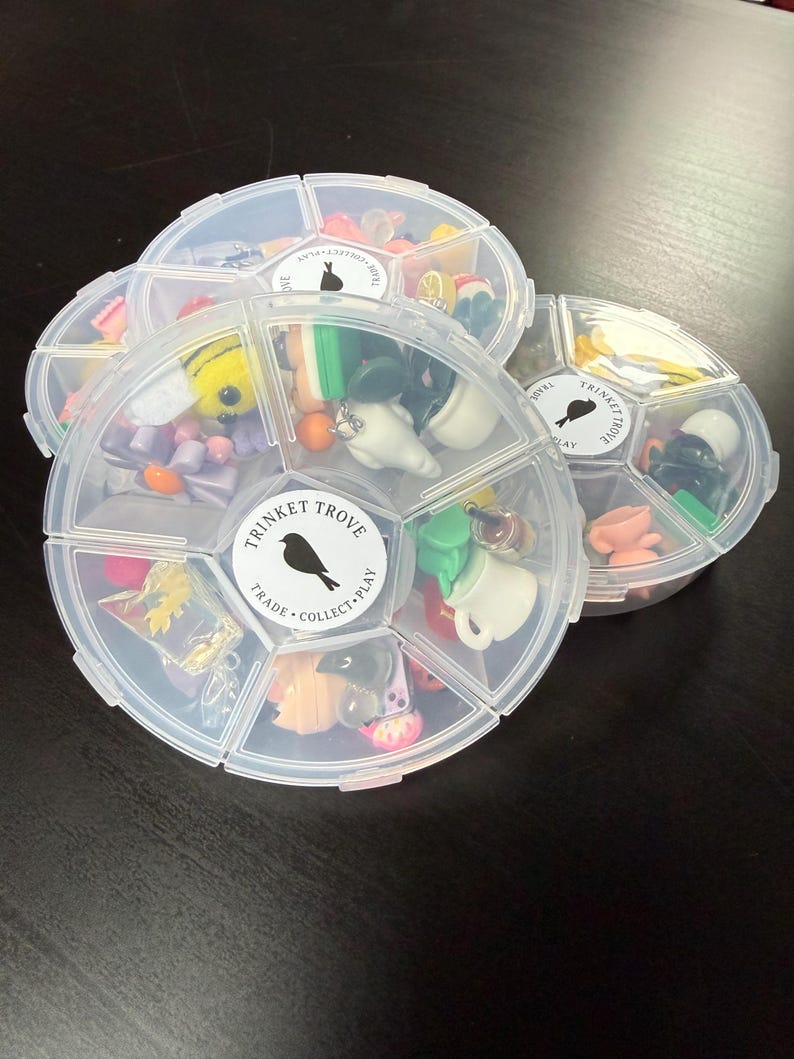 May include: Three clear, segmented containers filled with miniature trinkets. Each container has a label with a bird silhouette and the words "TRINKET TROVE, TRADE, COLLECT, PLAY." The trinkets include small figurines, beads, and charms in various colors.