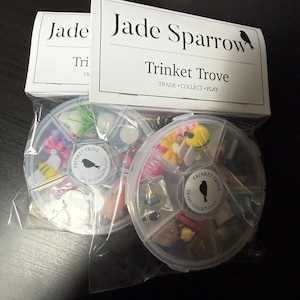 May include: Two clear plastic containers filled with small trinkets, each divided into sections. The containers are labeled "Trinket Trove" and "Jade Sparrow." The trinkets include miniature flowers, bees, and other small decorative items. The packaging reads "Trade, Collect, Play."