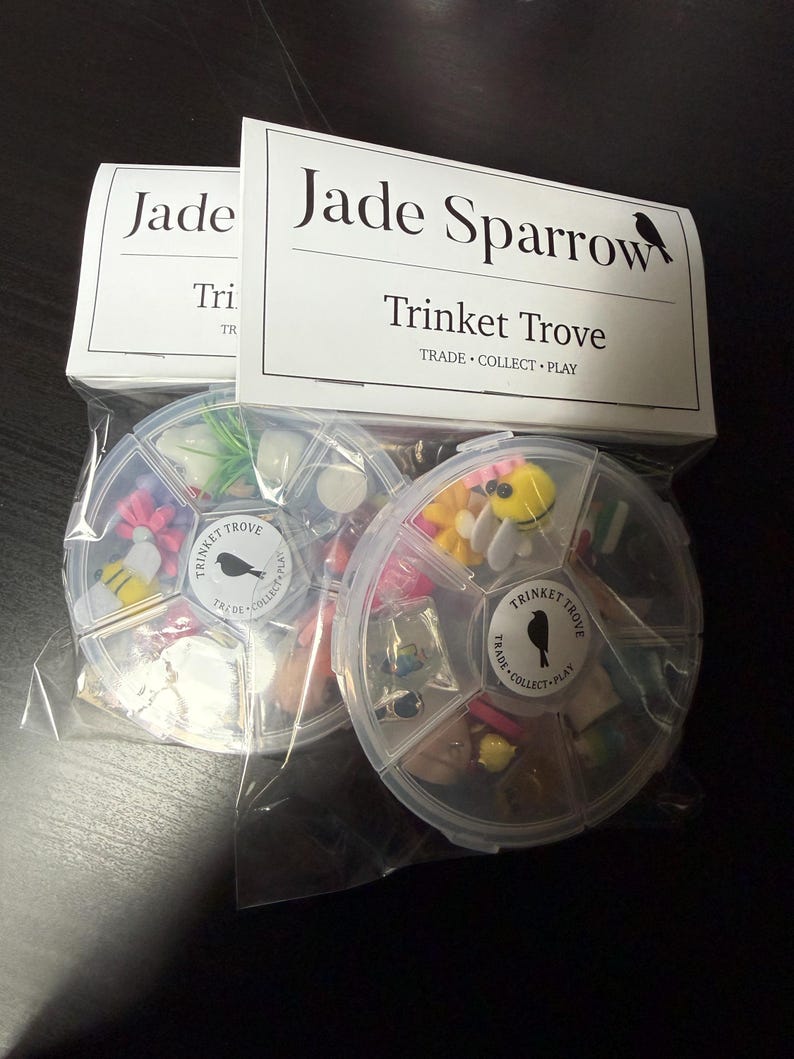 May include: Two clear plastic containers filled with small trinkets, each divided into sections. The containers are labeled "Trinket Trove" and "Jade Sparrow." The trinkets include miniature flowers, bees, and other small decorative items. The packaging reads "Trade, Collect, Play."