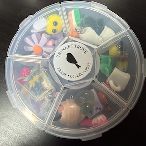 May include: A clear plastic organizer filled with various small trinkets. The organizer has a circular shape with multiple compartments. Trinkets include a yellow bee, a daisy, miniature food items, and other small charms. The center of the organizer has a label that reads "Trinket Trove".