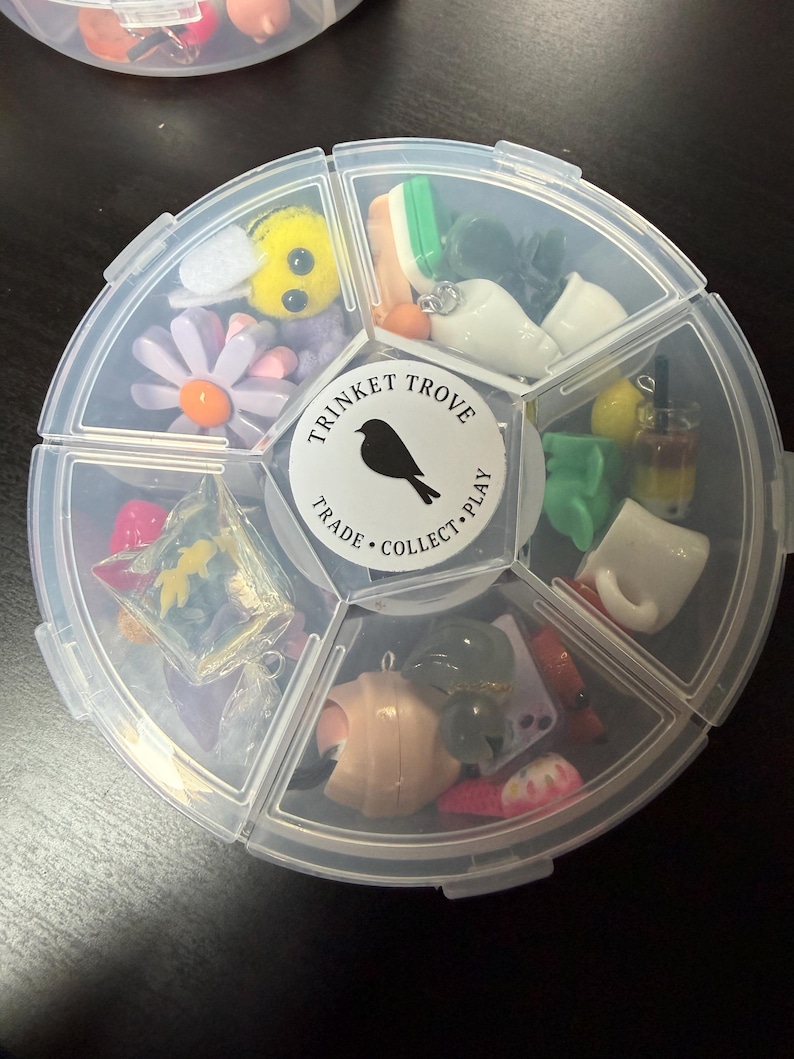 May include: A clear plastic organizer filled with various small trinkets. The organizer has a circular shape with multiple compartments. Trinkets include a yellow bee, a daisy, miniature food items, and other small charms. The center of the organizer has a label that reads "Trinket Trove".