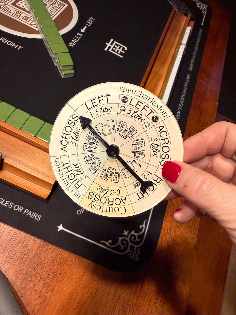May include: A circular Mahjong game direction indicator with a black arrow pointing to different directions. The indicator has text such as "Left", "Right", and "Across". The background includes Mahjong tiles and a wooden surface.