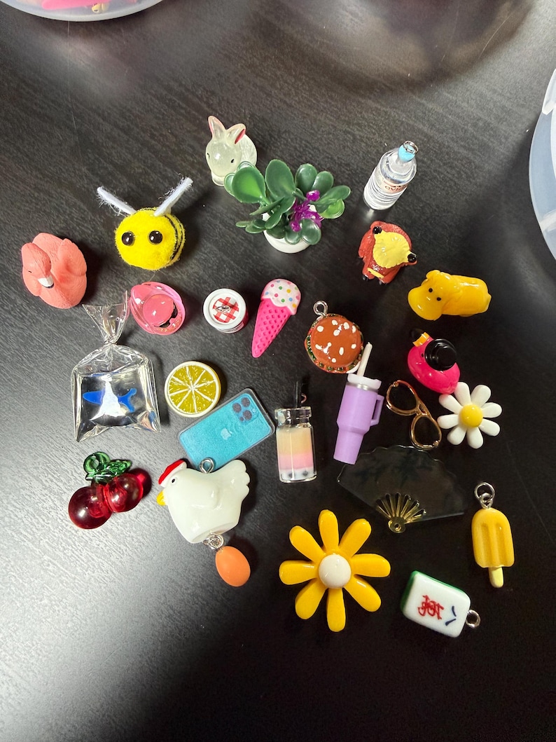 May include: A collection of colorful miniature charms, including a yellow bee, a pink ice cream cone, a miniature succulent plant, a yellow flower, and a miniature smartphone. Other charms include a burger, a chicken, and a boba tea.