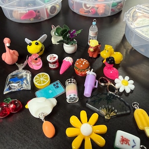 May include: A collection of miniature charms and trinkets, including a pink flamingo, a yellow bee, a miniature burger, and a yellow flower. The charms are stored in clear, divided plastic containers. Other charms include a miniature ice cream cone, a boba drink, and a miniature phone.