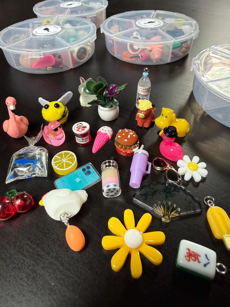 May include: A collection of miniature charms and trinkets, including a pink flamingo, a yellow bee, a miniature burger, and a yellow flower. The charms are stored in clear, divided plastic containers. Other charms include a miniature ice cream cone, a boba drink, and a miniature phone.